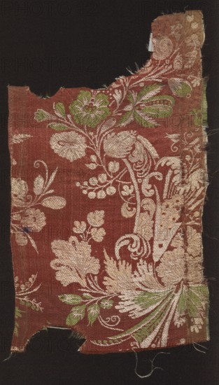 Fabric fragment. Fragments of silk patterned fabric with plant patterns; stripe decorated with colorful threads. Red satin background with motifs worked in green, white, and pink threads in a weft weave. Design consists of twigs with fanciful flowers of various species placed in cup-shaped forms; pattern size larger than preserved fragment. Motifs in the bizarre type of fabrics. Selvedge width: 0.4 cm red and white threads. Early 18th century, Italy. Height 36 cm, width 22 cm., A worn textile fragment with a dark background showing a floral damask pattern on a deep reddish-brown ground, featuring a group of pale beige ornamental leaves and flowers woven throughout and a few green leaves and floral accents; the fabric edges are frayed and irregular with some missing sections, and loose threads extend from the sides.