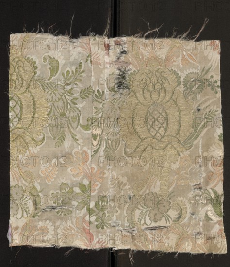Fabric fragment. Cream silk damask fabric, embroidered and brocaded with colorful threads. Promoting threads: green, light and dark salmon. Brooching thread: gold. Pale cream satin background with weft weave coverings, embroidery, and brocade. Fantastical floral decoration: small twigs and leaves of the damask sub-report complement larger motifs brocaded with gold threads. Stylized pomegranates pineapples placed on a winding stem, surrounded by sprigs of smaller flowers. Fabric sewn from two pieces. Composition of the report motifs based on mirror symmetry. Weave: satin weave with warp coverage; pattern creating a sub-report in a plain weave; colorful pattern promoted in zones; promoting threads tied in a twill weave introduced two at a time in the interlacing. Date: 1710–1715. Place of origin: France. Materials: silk, metal thread. Dimensions: height 33 cm, width 33.5 cm., A rectangular piece of fabric with frayed edges resting on a dark background shows an ornate floral and foliate w...