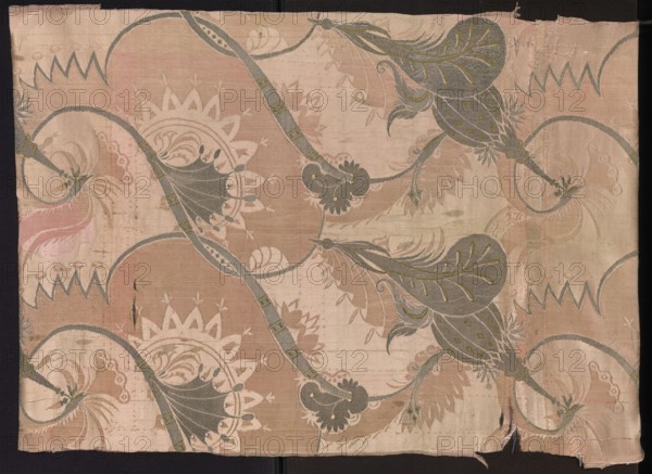 Fragment of pink silk brooch fabric with a fanciful and floral pattern in the Bizarr type. Early 18th century, France. Pattern: arrangement of intertwining tendrils with fanciful forms—some half-vegetal, half-animal fantastic winged dragons, others topped with sharp edges like poppy-head shafts. Construction: satin with warp cover and simple weave background; other motifs brocaded with metal threads. Colors: pink ground; brocades in silver, green, pale pink; silver metal thread with a loose braid. Width of fabric: 54.5 cm. Report dimensions: height 60 x width 27 cm; overall height 106 cm, width 54 cm., Classification: patterned silk fabric, fragment., A rectangular piece of patterned fabric with a faded background in shades of pale pink and beige, featuring a repeating ornamental design of flowing curved vines and large stylized leaves in muted gray-green with small olive accents. The fabric shows a few scalloped circular motifs in off-white and pale tan, and several areas of paler bei...