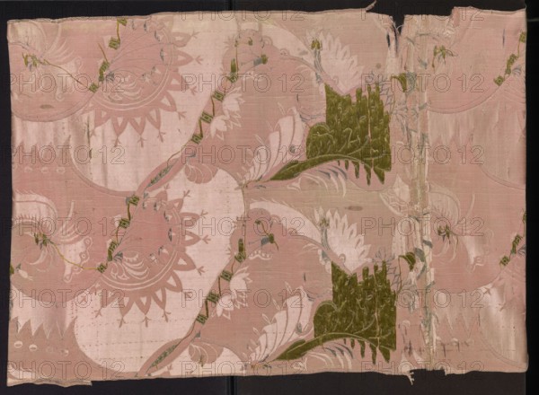 Fragment of pink silk brooch fabric with a fanciful and floral pattern in the Bizarr type. Patterned fabric fragment of silk, early 18th century, France. Motifs: arrangement of intertwining tendrils with fanciful forms — some half-vegetal, half-animal fantastic winged dragons, others topped with sharp edges like poppy-head shafts. Weave/technique: satin with warp cover and simple weave background; other motifs brocaded with metal threads; silver metal thread with a loose braid. Colors: pink background; brocades in silver, green, pale pink. Width of the fabric: 54.5 cm. Reported fragment dimensions: height 60 x width 27 cm; overall fabric height 106 cm, width 54 cm., A rectangular piece of patterned fabric displayed against a dark background, primarily in shades of pale pink and dusky rose with accents of olive green and muted gray; the fabric shows a repeating ornamental design of large, sweeping leaf- and petal-like shapes and curved scalloped motifs, with two prominent irregular oliv...