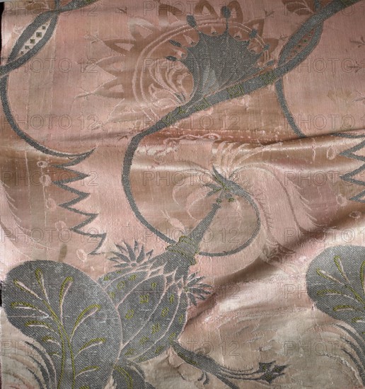 Fragment of pink silk brocade fabric with a fanciful floral pattern in the Bizarr style. Motifs: satin with warp cover and simple weave background; other motifs brocaded with metal threads. Pattern: intertwining tendrils with fanciful forms — some half-vegetal, half-animal fantastic winged dragons, others topped with sharp poppy-head–like shafts. Colors: pink ground; brocades in silver, green, pale pink, and silver metal thread with a loose braid. Width of the fabric: 54.5 cm. Reported dimensions: height 60 x width 27 cm; overall height 106 cm, width 54 cm. Early 18th century, France. Classification: patterned silk fabric, fragment., A single fabric panel with a flowing decorative pattern, showing a smooth, slightly shiny surface with visible weave texture and some creases. The design includes sweeping curved lines and stylized botanical shapes that resemble leaves and seed pods, with layered ornamental motifs and scalloped edges. Colors visible include a dominant soft pink background,...