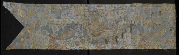 Fabric fragment, origin unknown, early 18th century, France. Silk lisère warp satin fragment cut in the form of a chasuble column with a triangular neckline; warp-woven lisère satin with prominent brocaded pattern. Right side: damask subrapport of lisère satin background and plain weaves decoration; left side: plain weaves. Decorated with whimsical plant motifs: tiny flowers on slender celadon-green branches and fanciful forms resembling onion-shaped flower buds growing from ornamentally treated horn-shaped bases; lace-like patterning. Colors: blue background with colorful brocade and embroidered threads. Brocade threads: gold, white, pale pink, blue, burgundy; embroidered threads: orange, pink, blue. Pattern created by threads tied in a plain/simple weave. Material: silk, lisère satin, embellished and brocaded. Dimensions: height 106 cm, width 23 cm; repeat report length 44 cm., A long horizontal piece of fabric with a pale blue background featuring an ornate floral and scroll pattern...