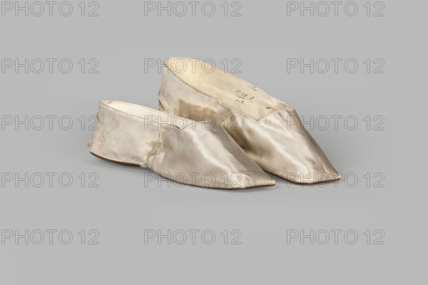 Women's slippers by Wincenty Witoszynski Warsaw; shoemaker's workshop; ca. 1840–ca. 1850. Shallow, flat women's slippers made of white silk. Symmetrical soles of thin leather with straight-cut toes. Lining: front of white canvas, back of thin white leather. Inside, an octagonal company label glued in: Wincenty Witoszynski / in Warsaw / at Senatorska Street next to the Main Town Hall No. 463. Upper part is machine-sewn. Date: 1840–1850, place of origin: Warsaw. Materials/typology: footwear, clothing. Dimensions: height 5.3 cm, width 5.3 cm, length 23.2 cm., A pair of pointed, slip-on shoes made of a shiny, smooth fabric in a pale ivory or cream color sit on a neutral light-gray background; the shoes show areas of slight discoloration and wear, with inner linings in an off-white or beige tone and slightly rougher fabric at the heels, and the soles appear to be a thin brown material visible along the edges.