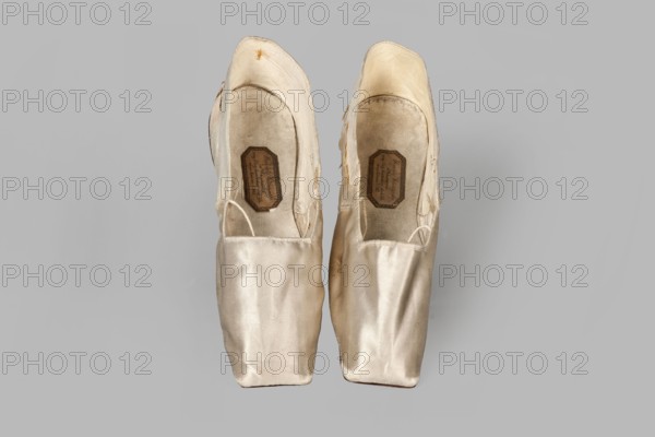 Women's slippers by Wincenty Witoszynski, ca. 1840–1850. Shallow, flat women's slippers made of white silk. Symmetrical thin leather soles with straight-cut toes. Lining: front of white canvas; back of thin white leather. Inside an octagonal company label: Wincenty Witoszynski / in Warsaw / at Senatorska Street next to the Main Town Hall No. 463. Upper part machine-sewn. Materials: silk, leather, canvas. Dimensions: height 5.3 cm; width 5.3 cm; length 23.2 cm., A pair of worn ballet pointe shoes seen from above, placed side by side on a neutral grey background; the shoes are a pale ivory to cream color with satin fabric showing creases and slight discoloration, the interiors appearing a bit darker with beige and tan tones, and there are visible areas where the fabric is frayed and stitched, along with a few straps of matching color looped inside.