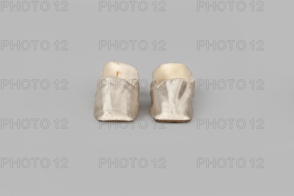 Women's slippers made of white silk with symmetrical thin leather soles and straight-cut toes. Lined at the front with white canvas and at the back with thin white leather. Inside is an octagonal company label reading Wincenty Witoszynski / in Warsaw / at Senatorska Street next to the Main Town Hall No. 463. Upper part machine-sewn. Shoemaker: Witoszynski, Wincenty; ca. 1840–1850, Warsaw. Function: women's footwear shallow, flat slippers. Materials: silk, leather, canvas. Dimensions: height 5.3 cm; width 5.3 cm; length 23.2 cm., A pair of small slip-on shoes placed side by side on a light gray background, made of a shiny pale fabric with a slightly crumpled texture, showing off-white inner linings and some areas of cream and beige tones, with the fabric edges appearing slightly frayed and a small darker stain visible on one inner lining, the soles appearing to be a light brown color.