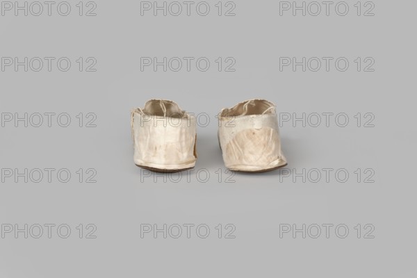 Women's slippers, made by Wincenty Witoszynski Warsaw; shoemaker's workshop; ca. 1840–ca. 1850. Shallow, flat women's slippers made of white silk. Symmetrical soles of thin leather with straight-cut toes. Lining of the front of white canvas, the back of thin white leather. Inside, an octagonal company label is glued in: Wincenty Witoszynski / in Warsaw / at Senatorska Street next to the Main Town Hall No. 463. The upper part is machine-sewn. 1840–1850. Place of origin: Warsaw. Category: footwear, clothing. Dimensions: height 5.3 cm, width 5.3 cm, length 23.2 cm., A pair of worn fabric shoes sits side by side on a plain surface, made of off-white cloth with visible creases and frayed edges, and showing areas of beige discoloration and light brown staining; the shoes have low, flat brown soles and slightly sagging openings, and the overall scene features a neutral light gray background and surface.