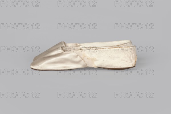 Women's slippers by Wincenty Witoszynski Warsaw; shoemaker's workshop; ca. 1840–ca. 1850. Shallow, flat women's slippers made of white silk. Symmetrical soles of thin leather with straight-cut toes. Lining of the front of white canvas, the back of thin white leather. Inside an octagonal company label is glued: Wincenty Witoszynski / in Warsaw / at Senatorska Street next to the Main Town Hall No. 463. The upper part is machine-sewn. 1840–1850. Origin: Warsaw. Materials: silk, leather, canvas. Function: women's footwear. Dimensions: height 5.3 cm; width 5.3 cm; length 23.2 cm., A pair of flat, slip-on shoes placed side by side on a plain background, made of a smooth, slightly shiny fabric with a pale cream color; the toe area appears slightly rounded and the uppers show some creasing and subtle discoloration in shades of beige and faint brown; the sides and inner lining display a softer, textured cream tone with a few worn areas and frayed edges, and the thin soles are a light brown visi...