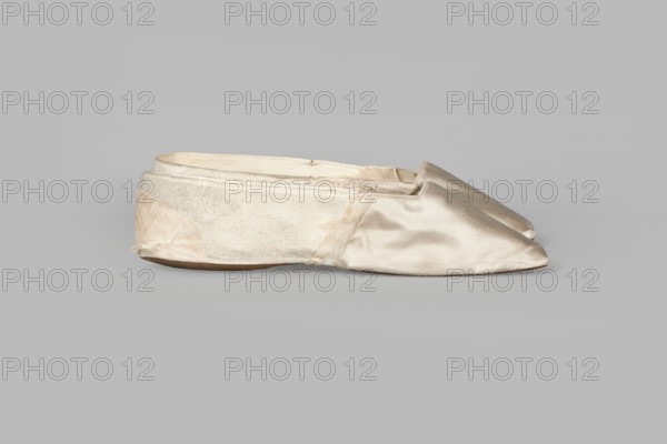Women's slippers by Wincenty Witoszynski, ca. 1840–1850. Shallow, flat women's slippers made of white silk. Symmetrical thin leather soles with straight-cut toes. Lining: front of white canvas, back of thin white leather. Inside an octagonal company label reads: Wincenty Witoszynski / in Warsaw / at Senatorska Street next to the Main Town Hall No. 463. Upper part is machine-sewn. Footwear; height 5.3 cm, width 5.3 cm, length 23.2 cm; Warsaw., A single worn ballet flat made of a shiny satin-like material rests on a plain background, showing frayed edges and areas of wear; the shoe appears light beige to off-white with slightly darker soiled patches, a satiny sheen on the toe area, and a matte textured rear section, with a thin brown sole visible underneath and subtle stitching along the seams.