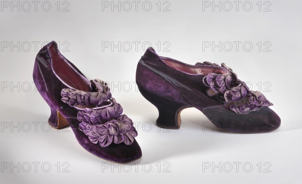 Women's slippers. Shallow slippers with two buttoned flaps at the front. Contoured heel. Upper made of dark purple velvet, machine- and hand-sewn. Front flaps decorated with purple ribbon draping. Lining at the toes made of white fabric; remaining lining pink glace leather. Endpaper of pink glace leather. No brand labels. Sole of unfinished leather, slightly rounded at the front. 1888, Poland. Footwear; women's footwear; height 12 cm, length 21.3 cm., A pair of purple velvet women's shoes with decorative purple fabric rosettes on the uppers, pinkish interiors, curved heels with brown undersides, and a plain light gray background.