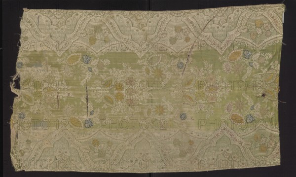 Fabric fragment, broached silk damask, early 1st quarter of the 18th century, France. Warp-covered satin ground with green warp. Broaching wefts in light and dark pink, pale and dark blue, celadon, yellow, and salmon; feature threads dark and light pink and white, captured in a weft weave. Unilateral two-color damask effect, with mirror symmetrical lace pattern: floral sprigs along a central axis and serrated lace border with floral bouquets. Stylized yellow and blue-green flowers on a light green background. Preserved selvedge, pile width, and pattern repeat. Pattern width 50 cm; repeat height 48 cm, repeat width 48 cm; edge width 0.6 cm. Fragment dimensions: height 79 cm, width 48 cm., A rectangular textile fragment with a faded pale green ground and an ornate woven border in light beige forming scalloped arches along the top and bottom edges, decorated overall with floral and foliate motifs; the surface shows several embroidered or woven flower clusters and leaves in muted yellow, p...