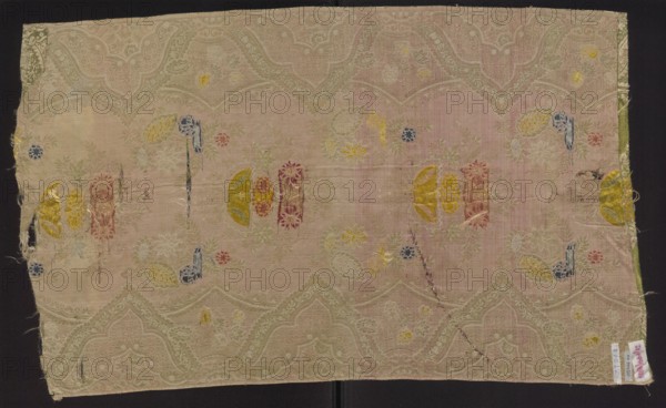 Fabric fragment, silk brocaded and broached damask, 1st quarter of the 18th century, France. Warp-covered satin ground with green warp. Broaching wefts in light and dark pink, pale and dark blue, celadon, yellow, and salmon; feature threads dark and light pink and white, captured in a weft weave. Unilateral two-color damask effect, broached with colored wefts. Lace-style motif arranged in mirror symmetrical composition: floral sprigs along a central axis and a serrated lace border with floral bouquets. Pattern width 50 cm; repeat height 48 cm, width 48 cm; edge width 0.6 cm. Fragment dimensions: height 79 cm, width 48 cm., A rectangular piece of woven fabric laid flat with a faded pink background and a subtle repeating scalloped pattern in a lighter beige tone, showing areas of wear and small holes. Scattered across the surface are embroidered or woven floral and abstract motifs in several colors including yellow, blue, red, green, and gold, with some motifs appearing as semi-circular ...