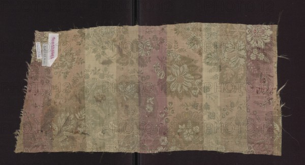 Fabric fragment, silk, early 18th century, France. Fragment of patterned fabric with preserved lace-like patterns and plant/floral motifs. Colors: green background with sand-beige and cherry weft. Decoration: large repeat with symmetrical, mirrored layout; repeat size 48.5 x 49 cm. Dimensions: height 52 cm, width 24.5 cm., A rectangular piece of woven fabric with frayed edges laid flat against a dark background, showing vertical bands of muted colors including beige, pink, green, and tan; the fabric surface features an allover floral and leafy woven pattern with various blossoms and foliage motifs in lighter and darker shades of the same palette, and areas of wear and discoloration; a small fabric tag is attached near one short edge; loose threads extend from the edges.