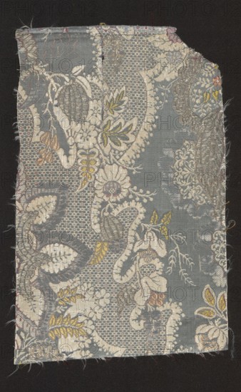 Fragment of fabric, fabrics, fragments of silk fabrics, patterned fabrics, fabrics with lace patterns, fancy motifs, floral motifs, plant motifs. Background - satin with a warp cover blue warps not tied in the fabric structure. Pattern - partially liseré with white threads - lace overlay. Colorful motifs brocaded with wefts: green, celadon, light and dark pink, salmon and silver. Promotion: with a dark cherry weft. Symmetrical report the preserved fragment is only half of the piece - lace motifs with a fine pattern imitating tulle eyelets and small flower sprigs. 1st quarter of the 18th century, France. Fabrics, height 32 cm, width 21 cm., A rectangular piece of woven fabric with frayed edges lies against a dark background, showing an ornate floral and leaf pattern in muted colors. The dominant background color is a soft gray-blue, overlaid with intricate designs in off-white and pale cream that form flowers, leaves, and decorative scrolls. Accents of muted yellow and olive appear on s...