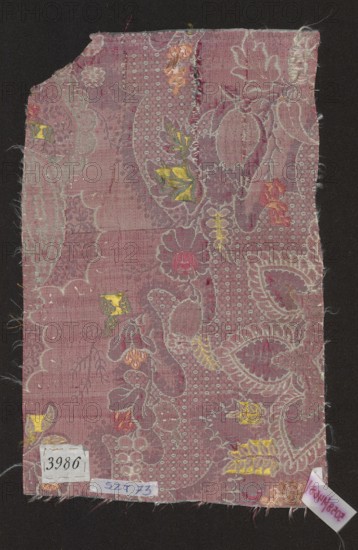 Fragment of fabric, fabrics, fragments of silk fabrics, patterned fabrics, fabrics with lace patterns, fancy motifs, floral motifs, plant motifs. Background - satin with a warp cover blue warps not tied in the fabric structure; pattern - partially liseré with white threads - lace overlay; colorful motifs brocaded with wefts: green, celadon, light and dark pink, salmon and silver. Promotion: with a dark cherry weft. Symmetrical repeat the preserved fragment is only half of the piece - lace motifs with a fine pattern imitating tulle eyelets and small flower sprigs. 1st quarter of the 18th century, France; fabrics; height 32 cm, width 21 cm., A rectangular textile fragment with frayed edges laid on a dark background, showing an overall mauve to dusty pink ground with a woven floral and paisley-like pattern in lighter pink and pale silvery threads, and small accents of yellow, gold, orange, and deeper pink. The fabric surface shows a few areas of wear and loose threads, and scattered small...