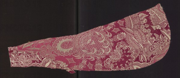 Fragment of fabric. Two fragments trimmed to the sides of a chasuble, single-sided damask; background: satin weave with a warp cover two warps — pink and white; decoration: white weft in plain and grosgrain weave. Pattern: white stylized flowers and fragments of larger lace patterns on a cherry-red background. Fragments of unsewn liturgical robe, patterned silk fabric, 17th century, France. Height 64 cm, width 22 cm., A curved textile fragment with an ornate floral and paisley pattern in shades of pink and cream set against a dark background, showing intricate woven motifs including stylized flowers, leaves, and circular medallions; the fabric edges are uneven and slightly frayed, and the pattern varies in density across the piece with areas of solid pink and areas where the cream weave forms detailed lace-like designs.