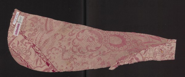 Fragment of fabric. Two fragments trimmed from the sides of a chasuble. Single-sided damask; background satin weave with a warp cover two warps — pink and white. Decoration executed with white weft in plain and grosgrain weave, featuring white stylized flowers and fragments of larger lace patterns on a cherry red background. 17th century, France. Fabrics; height 64 cm, width 22 cm., A single elongated fabric fragment with curved and straight edges lies against a dark background; the fabric shows an allover floral and ornamental woven pattern in shades of pink and lighter pink, with areas of deeper rose and pale blush creating the design; there are visible seams and stitched edges with some fraying and loose threads, and a narrow strip of contrasting patterned trim along one curved seam in darker pink and white; the dark background appears nearly black with a subtle central vertical seam.