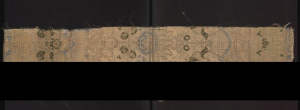 Fragment of fabric., Silk, 1st quarter of the 18th century, France. One-sided damask with floral and lace motifs; background satin with warp cover; pattern brocaded with colored threads. Symmetrical composition with axis at center of the piece. Large, ornate bouquets of flowers with heavy decorative heads on slender branches; flower leaves and parts of the background decorated with lace ornament poin de neige. Colors: salmon satin ground; brocade threads in green, white, and blue tied in rows. Height 95 cm, width 20 cm., A horizontal strip of patterned fabric with frayed edges sits against a dark background; the fabric shows an intricate floral and ornamental weave with a faded beige ground and accents of green and blue, with visible worn threads and small loose fibers along the top edge, a central seam or fold line running vertically, and areas where the weave appears slightly lighter or darker creating subtle texture variations.