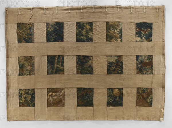 Tapestry — verdure;,  noted; late 17th century, Flanders. Subject: park/garden scene with animals and architecture. Composition: on the right, a fountain with two piled goblets, one supported by Atlas, with a dog and a deer in front; center foreground a bush with pumpkin fruit; background left garden architecture and center/right trees. Border with floral motifs, fruit, and musical instruments. Materials: fabrics. Colors: green, blue, brown, ochre, yellow. Dimensions: height 415 cm, width 297 cm., A rectangular textile composed of a plain beige-brown fabric formed into a grid with horizontal bands and vertical separators, revealing a group of darker decorative square panels set into the grid; the decorative panels show dense, textured imagery in deep green, dark blue, brown, and muted yellow tones with leafy and organic shapes, while the surrounding grid is a coarse, light tan-beige color with visible creases, stitched seams, and small fabric loops along the top edge.