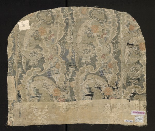 Fabric fragment, fabrics, fragments of a larger textile object, fragments of silk fabrics, patterned fabrics. Flaunted and brocaded silk. Damask subreport - background in a satin weave, pattern in a grosgrain weave, green flaunting thread tied in a twill weave. The white, green, and gold pattern provides a backdrop for colorful floral and fanciful motifs. The asymmetrical fabric report is created by larger forms resembling cornucopias, which intertwine turbinate, lace, and plant motifs. Bizarre decoration. Flaunting thread - green. Padding threads: salmon, pink, blue, gold braided thread. Beginning of the 18th century, France, fabrics. Dimensions: height 51 cm; width 61 cm; width of the brim 43 cm; width of the edge 0.3 cm; width of the report 23 cm., A worn rectangular textile fragment with an arched top displayed against a dark background, showing an ornate repeating paisley and floral design in muted colors including grayish blue, beige, pale green, soft orange, faded red, and hints...