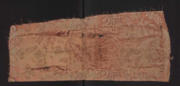 Fabric fragment, fragments of a larger textile object, patterned silk fabrics. Pale red pink lampas with a white patterned weft — a unilateral two-tone damask. Background: light red/pink warp satin; overlays in plain and liseré weaves with a white weft tied in a weft weft-faced weave; reverse side in plain weave. Repeat rapport composed of small flowers, fanciful leaves, and miniature lace motifs forming an ornament on the leaves; motifs typical of bizarre fabrics inspired by lace. Another fragment likely part of the same fabric shows a pale red stripe with a white pattern weft. Subreport: plain weaves pink warp/pink weft and white weft weaves. Colors: pink background with white decorative patterning. Hem width: 0.4 cm. Rapport: length 64.5 cm; width 24.5 cm. Overall fabric height 73 cm, width 31 cm. Early 18th century, France., A rectangular piece of worn fabric with a faded floral and paisley woven pattern rests against a dark background; the textile shows frayed edges and several lo...
