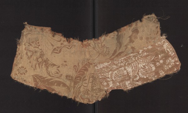 Patterned silk fabric fragment, early 18th century, France. Two-color damask with stylized leaves in a stripe weave structure; pale red warp satin background overlaid with plain and liseré weaves and accented by a white patterning weft tied in a weft weave; reverse in plain weave. Unilateral two-color damask; motifs of small flowers, fanciful leaves, and miniature lace-inspired motifs forming ornaments on the leaves—patterns in a bizarre fabric type inspired by lace. Repeat dimensions exceed the preserved fragment. Hem width: 0.5 cm. Dimensions: height 53 cm, width 19 cm. Fabric fragment no. SZT 78 is probably part of the same fabric., A worn, torn fabric fragment with a floral and paisley woven pattern lies against a dark background; the textile shows frayed edges and irregular cuts, with decorative motifs including stylized flowers and curved leaf shapes visible across its surface. The colors visible are shades of beige, tan, brown, and a lighter pale metallic tone that appears as a ...