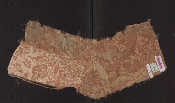 Fabric fragment, Patterned silk in a two-color damask type with a decoration of stylized leaves. Stripe weave structure, pale red accented with a white patterning weft. A type of unilateral two-color damask. Background — light red warp satin, overlaid with plain and liseré weaves, accented with a white weft tied in a weft weave; reverse side in a plain weave. The repeat is composed of small flowers, fanciful leaves, and miniature lace motifs forming an ornament on the leaves. The repeat dimensions are larger than the preserved fragment. Motifs in the bizarre fabric type inspired by lace. Fabric fragment no. SZT 78 is probably part of the same fabric. Hem width: 0.5 cm. Early 18th century, France. Height 53 cm, width 19 cm. Materials: silk. Classification: fabrics, fragments of patterned silk., A worn textile fragment with a decorative floral and paisley pattern sits against a dark background; the fabric shows frayed edges and loose threads and has a sheen that highlights its woven moti...