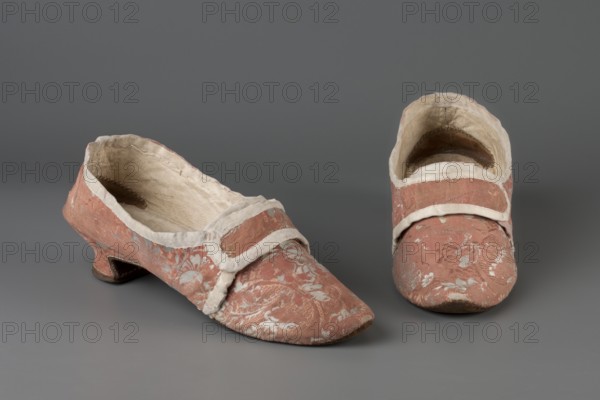 A pair of women's slippers made of patterned silk damask lined with leather. Dark pink outer fabric with a white floral motif. Shallow slippers, fitted with two decorative straps at the front. 3.5 cm heel, tapering down from mid-height. Symmetrical soles, pointed towards the front. Fabric-covered heel. Hand-sewn. Toes lined with canvas; heel lined with leather. Endpapers made of thin white leather. 2nd half of the 18th century, Europe. Height 9 cm, length 25 cm., A pair of low-heeled shoes sit on a neutral gray background, both covered in a faded pink fabric with a floral woven pattern in a lighter color; the shoes have pale off-white trim around the openings and across the insteps, and the interiors show a worn beige lining; one shoe is shown from an angled side view revealing a short curved heel and scuffed sole, while the other is shown from the front with a rounded toe and visible interior shadowing.