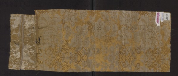 Fabric fragment, fragments of silk fabric, patterned two-color damask in olive and white. Weave construction: two-color warps olive and white; background a derivative of plain weave olive with sub-ornament promoted by olive warp threads and sparse weft interlacing a type of grodetur. Pattern: derivative of plain weave white wefts and white nooses. Decoration: symmetrical, mirror-like arrangement with axis of symmetry at center of panel; branches of stylized flowers dominated by large blooming peonies; sub-ornament features floral and lace motifs. Colors: olive background, white motifs. Wefts: white and olive. Warps: white and olive; leading—olive. Measurements reported: height 40 cm; also recorded height 65 cm, width 24 cm; edge width 0.4. Date: 1st half of the 18th century. Probable origin: France., A rectangular piece of woven fabric with a floral damask pattern lying on a dark background, showing a larger main panel and a narrower attached strip on the left; the fabric displays a me...