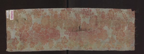 Fabric fragment, fragments of silk fabric, patterned two-color damask, 1st half of the 18th century, France . Salmon-pink plain-woven background with pearly-blue decoration. Subornament formed by released warp thread in background color; in the pattern area the subornament drawing is not connected to the wefts by any weave. Pattern: winding lace ribbon subornament, large and small flowers on winding stems peonies or roses. Two warp colors: pink and blue; two weft colors: pink and blue. Piece height 55 cm report, another measurement given as height 74 cm, width 24 cm; edge 0.5 cm. Similar pattern appears in dress fabric no. SZT 2745 and fragments SZT 80, SZT 60. Materials: silk. Function: fabric fragment., A rectangular fabric panel with a floral woven pattern laid against a dark background, showing a repeating motif of large and small stylized flowers and leaves. The fabric is primarily light with a pale greenish or silvery ground and a coral to muted pink floral design, with some area...
