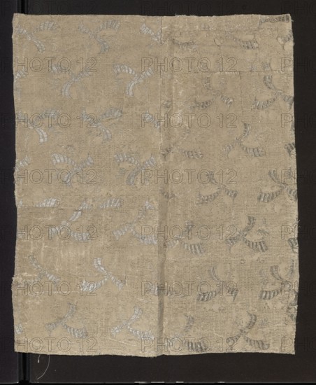 Fabric fragment, fragments of silk patterned fabric with preserved pattern. Fragment showing a pattern of leafy twigs tied with a kind of ribbon, arranged in rows with shifts and changes of direction. Colors: white and silver background, silver motifs. Pattern size: 12 x 8.7 cm. 17th century, France. Fabric height 46.5 cm, width 39 cm., A rectangular piece of woven fabric with a beige ground and a repeating decorative motif in a pale silvery gray, laid flat on a dark background; the fabric shows a visible vertical seam a bit off center, some puckering and worn areas, raw fraying edges on the sides, and a textured surface with woven horizontal lines and subtle sheen where the pale silvery gray motif catches the light.