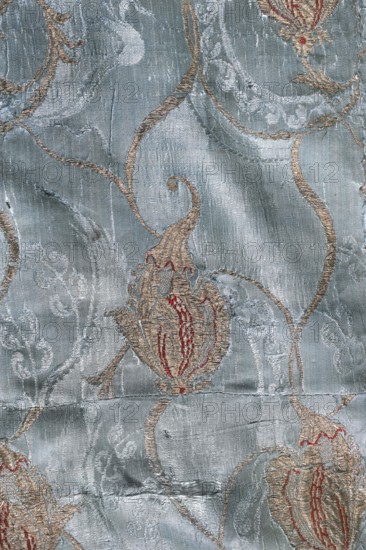 Fabric fragment, patterned silk fabrics, fragments of silk damask, pale blue ground brocaded with metal thread in a Bizarre type double-sided. Design in a net-and-strip arrangement with fanciful floral brocaded forms, partially outlined with a sinuous S-shaped brocaded gold line repeated across the report. Motifs include small twigs with unfolded leaves, winged forms, and shell-buds. Colors: blue background, gold and red stitching. Structure: background warp satin, sub-stitching plain weave, stitching with wefts gold and red. Edge width: 0.5 cm. Report width: 17.5 cm. Ca. 1712–1720, France. Overall dimensions: height 126 cm, width 85 cm., A close-up of a patterned fabric showing a textured, slightly shiny background in shades of light blue and silvery blue with a few raised, curving embroidered designs in beige and tan; there are accents of muted red within some floral-shaped embroidered motifs, and the surface shows subtle creases and weave irregularities.