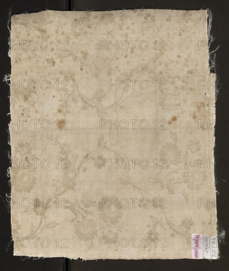 Fabric fragment, silk, patterned floral double-warp fabric. Cream and silver lamé with a pattern of twisting floral branches. 18th century, France; height 47 cm, width 39 cm., A square piece of fabric with frayed edges shown against a dark background, the fabric is an off-white to beige color with a subtle floral damask pattern visible across its surface; there are several brownish stains and spots scattered on the fabric and a faint horizontal fold line across the middle; a small rectangular tag is attached near one corner with a hint of pink and blue visible on it.