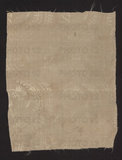 Fragment of silk fabric, damask with plain weave background and a lisere pattern, floral pattern, 18th century, Europe; dimensions: 12.5 x 19 report and height 29 cm, width 23 cm., A rectangular piece of light beige fabric with a subtle woven floral and paisley pattern is laid against a dark black background; the fabric shows frayed edges at the top and bottom and uneven trimming at the sides, with faint variations in beige tone where the weave and pattern catch the light, and a few tiny dark specks and slightly darker spots visible on the surface.