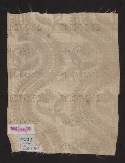 Fragment of silk fabric, patterned floral damask with plain weave background and lisere pattern; 18th century, Europe. Dimensions reported 12.5 x 19 cm also noted height 29 cm, width 23 cm., A rectangular piece of woven fabric with a subtle damask pattern featuring flowing paisley and floral motifs in varying shades of beige and cream, placed against a dark black background; the fabric shows frayed edges at the top and bottom, slight creases and small scattered darker spots, and a few small paper labels affixed near one corner whose printed content is not described.