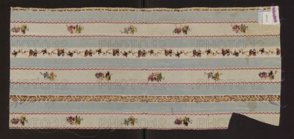 Fragment of silk fabric with a pattern of alternating vertical stripes of blue and cream; colorful motifs of small flower sprigs and tiny floral tendrils on the light stripes. 19th century, France. Silk fabric; height 72 cm, width 32.5 cm., A rectangular piece of fabric displayed against a dark background shows horizontal bands in alternating pale blue and off-white tones with decorative narrow borders in pink and red; scattered across the off-white bands are small floral-like motifs in shades of green, yellow, pink, and brown; a central off-white band contains a continuous embroidered vine-like pattern in brown and dark rust colors; along one edge there is a braided trim combining yellow, brown, and pink threads with some loose ends; the fabric edges are slightly irregular with a corner cutaway and some fraying, and overall the visible colors include pale blue, off-white, pink, red, green, yellow, brown, and rust.