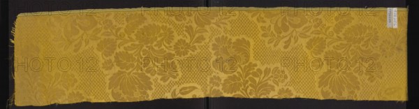 Fabric fragment, silk damask fragment, patterned floral motifs, preserved selvedge. Yellow damask with floral bouquet motifs on a background filled with a zigzag ornament. Background woven in warp satin 7/1; pattern executed in gros de Tours. Motif height 47 cm; selvedge width 0.8 cm threads: white, yellow, green. Overall fragment height reported 47 cm. Ca. mid-18th century, possibly France. Reported fabric dimensions: height 101 cm, width 20 cm., A long rectangular textile sample with a floral damask pattern sits against a dark background; the fabric is predominantly golden yellow with areas of slightly darker yellow forming large stylized flower and leaf motifs and textured chevron-like fields, and the edges show frayed threads on one side and a finished narrow hem on the opposite side.