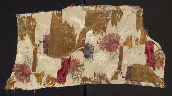 Fragment of upholstery fabric, silk and cotton brocade with cream grosgrain plain-weave background, brocaded in colorful threads and metal threads. Floral and fantastic motifs: winding twigs with fanciful flowers and plumes of leaves resembling bird wings; patterned fabric with preserved selvedge. Colors: red, dark and light pink, dark and light brown, damp green, blue, silver metal thread, copper metal thread. Dimensions: height approx. 64–71 cm, width 32 cm; edge width 0.9 cm. Possibly French, 18th century ca. 1760–1777 or early 19th century influence., A rectangular piece of fabric with frayed edges laying on a dark background, showing a patterned surface with floral and abstract woven motifs in shades of cream, beige, brown, gold, rust, red, pink, purple, gray, and green; several clustered flower-like shapes and leaf forms are woven into the cloth, along with textured vertical and curved bands that appear distressed or worn in places, and a few loose threads visible around the bord...