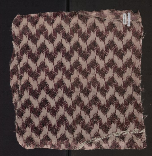 Fabric fragment of patterned silk with floral motifs on a white background motifs in pale pink, plum, and black. 2nd half of the 18th century, France. Height 51 cm, width 48 cm., A square piece of frayed fabric with a repeating geometric floral-like pattern in shades of mauve, burgundy, dark brown, and black on a pale pink background, laid against a dark surface; the fabric edges are worn and uneven with some loose threads and a narrow folded hem along one edge, and a small curved strip of lighter material with a faint spotted design is visible near one corner.