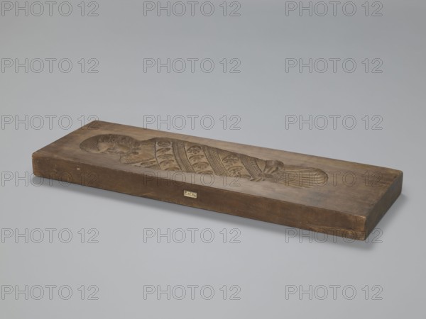 Double-sided gingerbread mould. Side A: image of a bachelor; Side B: baby in swaddling clothes. Maker unknown woodcarver or gingerbread maker. Double-sided, rectangular mould. One side shows a man in a long wig and tricorn hat, dressed in a costume probably dating to the turn of the 17th and 18th centuries: justaucorps with decorative cuffs, culottes, stockings, and high-heeled shoes; at his feet floral and geometric decorative motifs and the date 1827. Other side: baby in swaddling clothes richly decorated with geometric and plant ornament. Inscription on the edge: FW FIEBIGER. a. BAUTZEN. 17 signature of the manufacturer and beginning of the date. Materials/genre: gingerbread mould, woodcarver/gingerbread maker; style: folk motifs, plant ornaments, floral decorations, historical costume; region/date: Silesia Prussia, 1827. Dimensions: height 64 cm, width 22.5 cm, depth 4.3 cm., A rectangular wooden block in brown rests on a flat light gray surface against a pale gray background, the ...