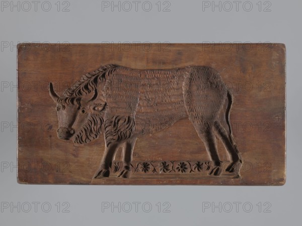 Double-sided gingerbread mould. Side A: bison; Side B: three representations — a bachelor in early 19th-century costume hat, justokor, artificial trousers, high boots, a baby in swaddling clothes decorated with a plant twig, and a fish with an elongated, scaled body. Rectangular wooden mould with the date 1806 engraved on the edge; the middle of the date shows the letter S against an anchor manufacturer/gingerbread maker mark. Materials: wood; function: gingerbread mould; style/subject: figurative scenes, human figures, zoomorphic motifs, folk art, historical costume; dimensions: height 19.5 cm, width 10.5 cm, depth 4.3 cm. Place/period: Silesia, 1806., woodcarver or gingerbread maker., A rectangular wooden panel with a carved relief of an animal spanning most of its surface; the carving depicts a shaggy-bodied creature with curved horns, a textured coat, a mane of flowing carved lines, and visible legs and hooves standing on a decorated base with carved floral motifs. The wood shows r...