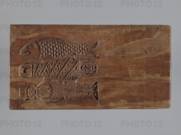 Double-sided gingerbread mould. Side A: bison; Side B: three figures — a bachelor in early-19th-century costume with a hat, wearing a justokor, artificial trousers and high boots; a baby in swaddling clothes decorated with a plant twig; and a fish with an elongated, scaled body. Rectangular wooden mould with an engraved date 1806 on the edge; the middle of the date shows the letter S against an anchor manufacturer's/gingerbread maker's mark. Materials: wood; function: gingerbread mould; style: folk art/folk customs; iconography includes Striding Bull, figurative and zoomorphic motifs, decorative art. Date: 1806. Place: Province of Silesia 1742–1918. Dimensions: height 19.5 cm, width 10.5 cm, depth 4.3 cm. Maker: unknown woodcarver or gingerbread maker., A rectangular wooden board with a smooth brown surface shows carved reliefs of a fish at the top, a patterned wrapped figure in the middle, and a standing humanlike figure at the bottom; the carvings show textured scale details on the f...
