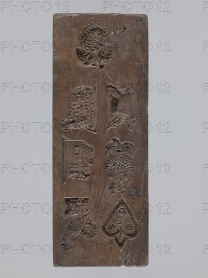 Double-sided gingerbread mold; obverse: seven different motifs — a heart, a rooster, a dog in front of a kennel, a woman and a man in costumes from the second half of the 19th century, a wild boar, a lamb and a fish; reverse: a fish covered with scales. 19th century, Province of Silesia 1742–1918. Height 28.5 cm, width 10.7 cm, depth 5.5 cm., A rectangular wooden board with a vertical orientation showing carved recessed motifs arranged in two vertical columns; the wood is brown with variations in tone and a weathered, slightly worn surface; a carved flower with layered petals and leaves appears near the top, and below it are carved animal and object shapes including a textured sheep-like head, a reclining animal, a house-like form, a bird with patterned feathers, a draped textile or flowing mane, and a heart-shaped spade motif with decorative inner carving; the carvings show tool marks and varying depths, and the board has small holes, surface cracks, and areas of darker brown patina.