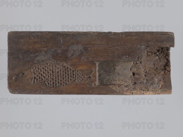 Double-sided gingerbread mold; obverse: seven motifs — a heart, a rooster, a dog in front of a kennel, a woman and a man in 19th-century costume, a wild boar, a lamb, and a fish; reverse: an image of a fish covered with scales. 19th century, Province of Silesia; height 28.5 cm, width 10.7 cm, depth 5.5 cm., A rectangular wooden plank with a carved fish motif in relief, showing detailed overlapping scales and a visible head and tail, with a worn and rough texture across the surface; one end of the plank appears damaged and crumbled with irregular holes and a fragmented edge, while the rest shows tool marks, indentations, and small circular blemishes; colors visible include shades of brown on the wood, darker brown in carved recesses, and patches of gray and muted ochre on the damaged area, all set against a pale gray background.