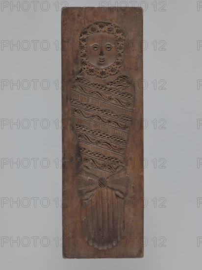 Double-sided gingerbread mould. Side A: soldier in parade uniform from the early 19th century—high shako with tassel, epaulettes, decorative sleeves, an order pinned to the chest, sabre with decorative tassel; geometric and plant decoration at the bottom. Side B: baby in richly decorated swaddling clothes with geometric and plant ornament. Rectangular wooden mould, engraved on the edge with the letter B initial of the maker. 19th century; Silesia Prussia; 1742–1918. Form. Height 35.3 cm; width 11.6 cm; depth 3.5 cm. Materials/technique: wood gingerbread mould; folk motifs, decorative art., unknown provenance., A tall rectangular wooden panel carved in brown wood features a stylized human figure in relief; the figure has a round face framed by a floral halo-like pattern, simple carved eyes, nose, and mouth, and a body wrapped in an intricately patterned shroud with diagonal bands of carved motifs, ending in a tied knot and a scalloped base; the background around the figure is the same b...