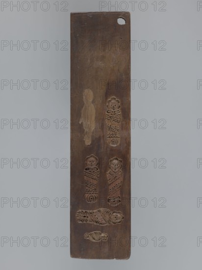 Double-sided gingerbread mould. Obverse: four images of human figures and plant motifs; Reverse: five images of infants of various sizes. Rectangular double-sided mould; one side: at the top a female figure in a long patterned dress with a low neckline, below a basket of flowers, below that a seated young naked man with long hair sitting among flowers and smoking a pipe, at the feet of the figure a skull with crossbones and the inscription MEMENTO MORI, and at the very bottom a wreath woven from leaves and flowers. The other side: five images of infants in patterned swaddling clothes. Gingerbread mould, decorative folk motifs, wreaths, female figures in rich dresses, Transience personification, floral decorations, allegorical representations, infants. 19th century, Province of Silesia 1742–1918. Form: rectangular; height 36.5 cm, width 8.8 cm, depth 1.5 cm., A long rectangular wooden board with a brown surface and a rounded top corner showing a small hole, set against a light gray back...