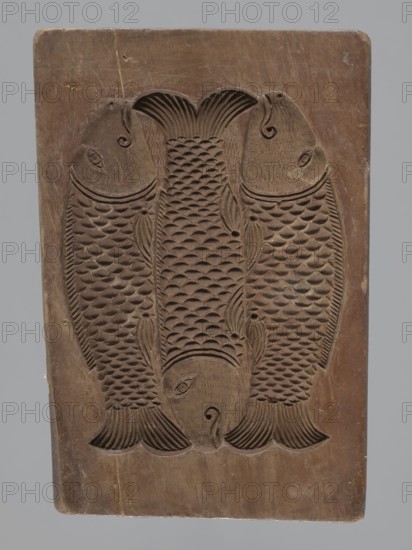 Double-sided gingerbread mould. Obverse: group of musicians; reverse: three fish. Double-sided, rectangular form; on one side a group of three musicians in costumes typical of the 17th–18th centuries—hat, justaucorps, culottes and stockings—first playing double bass, second trumpet, third violin, with floral and plant decoration at the bottom. Reverse: three stylized elongated fish covered with scales, shown in profile, arranged in a row. Gingerbread mould; decorative art; folk motifs, plant ornaments, zoomorphic motifs; early 19th century, Province of Silesia 1742–1918. Height 21.5 cm, width 15 cm, depth 2.8 cm., A carved wooden rectangular block with a relief of a group of fish arranged symmetrically, showing carved scales, fins, and eyes; the wood surface has a worn, smooth appearance with visible tool marks and small surface scratches; colors visible include shades of brown in the wood and a neutral gray background.