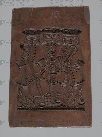 Double-sided gingerbread mould. Obverse: group of musicians; Reverse: three fish. Double-sided, rectangular form; on one side a group of three musicians in costumes typical of the 17th–18th centuries—hat, justaucorps, culottes and stockings—the first playing a double bass, the second a trumpet and the third a violin; floral and plant decoration at the bottom. Reverse: three stylized elongated fish covered with scales, shown in profile and arranged in a row. Gingerbread mould; decorative art; folk motifs; zoomorphic motifs; floral decorations. Early 19th century, Province of Silesia 1742–1918. Height 21.5 cm, width 15 cm, depth 2.8 cm., A rectangular wooden panel with a carved scene of a group of three standing figures wearing tricorn-style hats and long coats, each figure holding a stringed instrument and shown with detailed facial features and curled hair; the carving includes decorative carved foliage at the figures' feet and patterned borders near the bottom. The wood is a medium to...