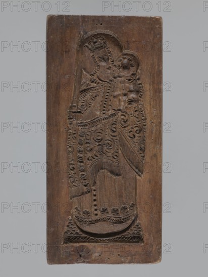Double-sided gingerbread mould. Obverse: image of Madonna and Child. Reverse: small image of Madonna in oval. Double-sided, rectangular form; on one side a figure of Madonna and Child, with a crown on her head, in a long patterned robe decorated with various geometric and plant motifs; the Madonna holds the Child in her left hand and a sword in her right. The other side: a smaller figure of the Madonna in an oval, surrounded by rays, the whole surrounded by a laurel wreath. Gingerbread mould; decorative folk motifs; religious motifs; Madonna and Child iconography; crown and sword iconography. 19th century, Province of Silesia 1742–1918. Height 25.8 cm, width 12 cm, depth 4.5 cm., A vertical rectangular wooden panel with a carved raised design showing a robed figure holding a smaller robed figure, both rendered in shallow relief with patterned decorative carving; the wood is brown with variations in tone and visible grain, and the background behind the panel is a uniform light gray.