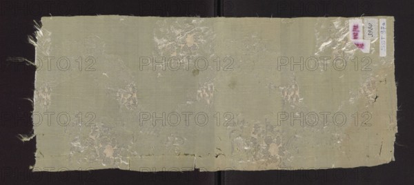 Fabric fragment, fabrics, fragments of a larger textile object, patterned silk fabric fragment, fragments of silk fabrics, floral patterns. Fragment of a green silk fabric with a delicate white lace report featuring a ribbon and floral sprig motif. This type of pattern with a graphic design appeared in the 1760s. The lack of gold and silver threads indicates that this fabric was intended for everyday clothing. The light green background and subtle pattern are characteristic of the fashion of the second half of the 18th century. Fabrics for dresses became lighter and their colors brighter. Colors: vert pomme apple green and vert celadon celadon green. Hem width: 0.3 cm. Density: 47 wefts, 76 warps. Date: 1760–1765. Place of origin: France. Height 56 cm, width 23 cm., A rectangular piece of frayed fabric laid against a dark background, showing a woven texture with irregular edges and some loose fibers; the fabric is primarily a muted pale green with areas of lighter and darker green shad...