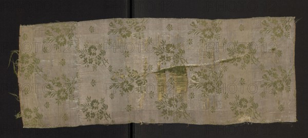Fragment of a solid silk fabric, patterned, with preserved floral motifs: two joined branches with unfolded roses and small flowers arranged in an alternating rhythm. Dimensions of fragment: 16 x 11.5 cm reported; overall height 61 cm, width 23 cm. Date: second half of the 18th century. Possible origin: Poland, Grodno., Classification: patterned silk fabric, fabric fragment., noted., A rectangular piece of woven fabric with a worn, frayed edge laid flat against a dark background, showing a repeating floral motif with stems and leaves in green on a pale beige or off-white ground; areas of the fabric show visible wear, thin spots, and a torn section with loose threads and exposed warp and weft in yellowish and greenish tones, and there are faint shiny threads that catch the light creating subtle silvery highlights across the surface.